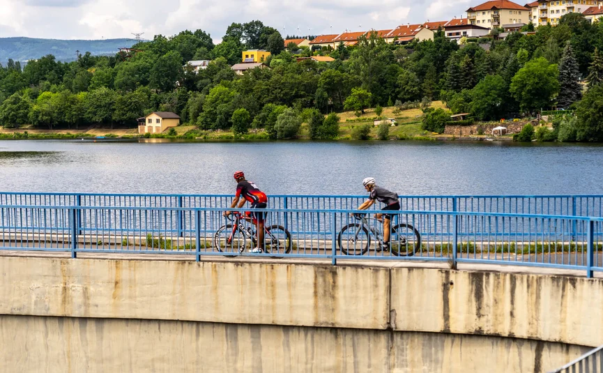 Zwei Radfahrer auf einer Brücke über einem See, Häuser im Hintergrund
