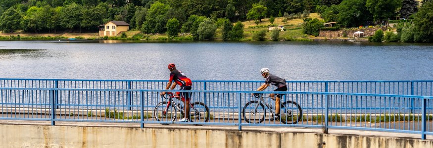 Due ciclisti su un ponte sopra un lago, case e alberi sullo sfondo