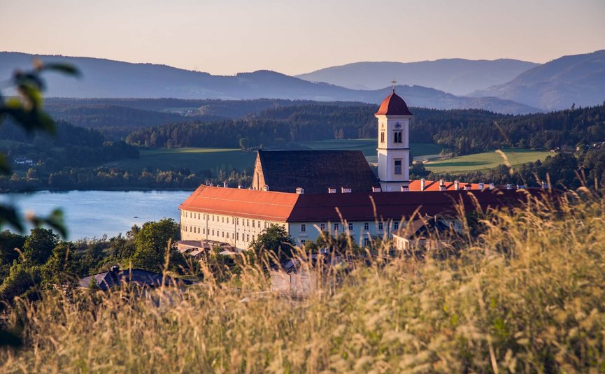 Stift St. Georgen **** © Elias Jerusalem Klostergebäude mit rotgedecktem Turm vor einem See und Bergen bei Sonnenuntergang