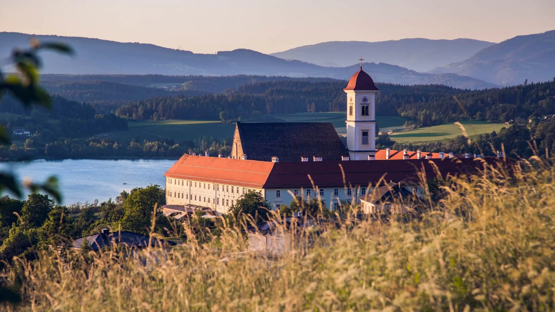 Klostergebäude mit rotgedecktem Turm vor einem See und Bergen bei Sonnenuntergang