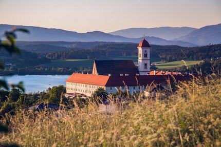 Stift St. Georgen © Elias Jerusalem Klostergebäude mit rotgedecktem Turm vor einem See und Bergen bei Sonnenuntergang