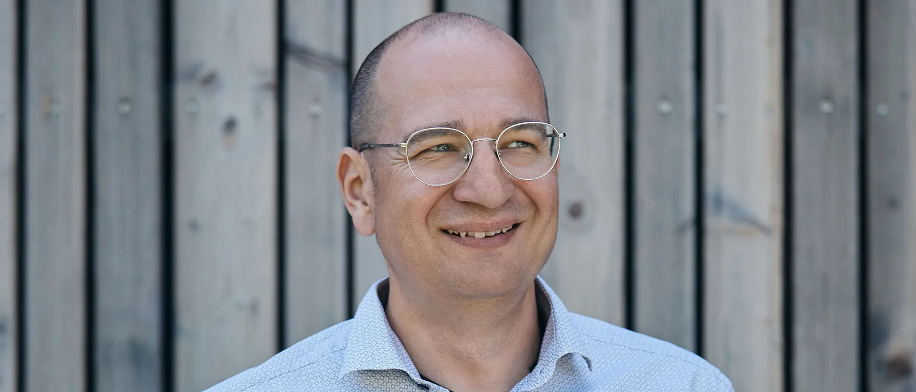 Lächelnder Mann mit Brille vor Holzwand, Hände in den Hosentaschen