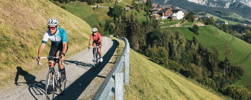 San Vigilio-Pässe-Quartett © Alex Moling Zwei Radfahrer fahren auf Bergstraße mit Dorf und Bergen im Hintergrund