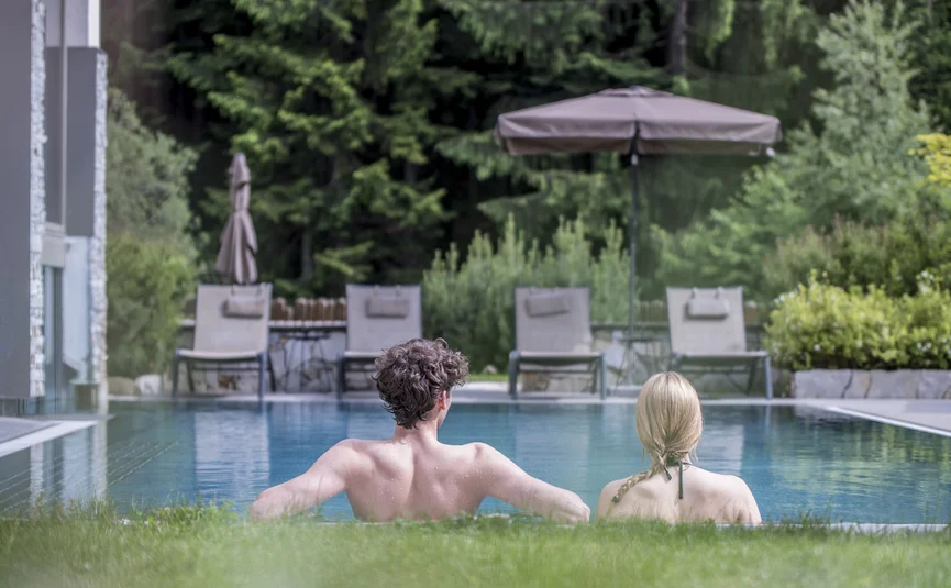 Paar entspannt im Pool mit Liegestühlen und Sonnenschirm im Hintergrund