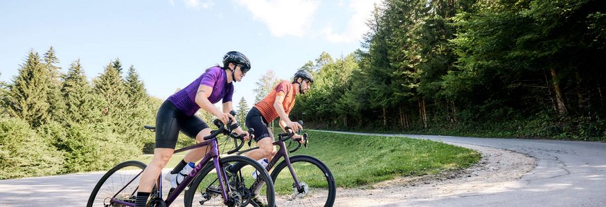 Zwei Radfahrer auf der Straße in einer bergigen Waldlandschaft.