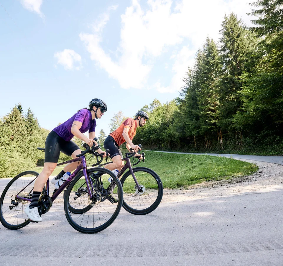 Zwei Radfahrer auf der Straße in einer bergigen Waldlandschaft.
