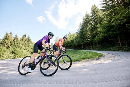 Zwei Radfahrer auf der Straße in einer bergigen Waldlandschaft.