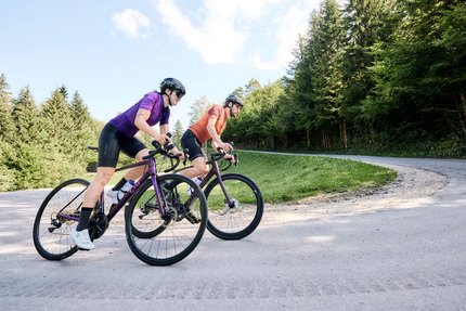 Zwei Radfahrer auf der Straße in einer bergigen Waldlandschaft.