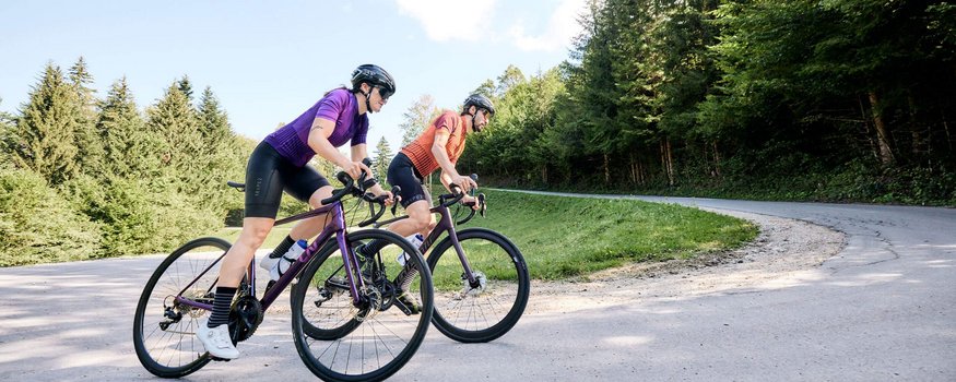 Zwei Radfahrer auf der Straße in einer bergigen Waldlandschaft.