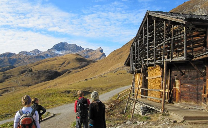 The Mountains are calling Paket Wandelaars op bergpad naast rustiek houten huis met bergen op de achtergrond