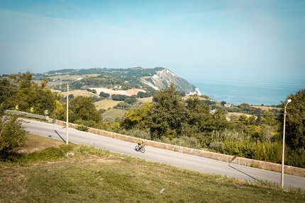 Ciclista su strada con colline e mare sullo sfondo in una giornata limpida