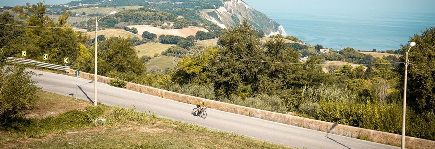 Ciclista su strada con colline e mare sullo sfondo in una giornata limpida
