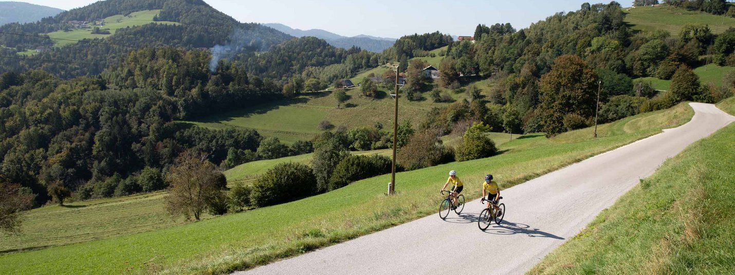 Zwei Radfahrer auf Landstraße mit grüner Hügellandschaft im Hintergrund