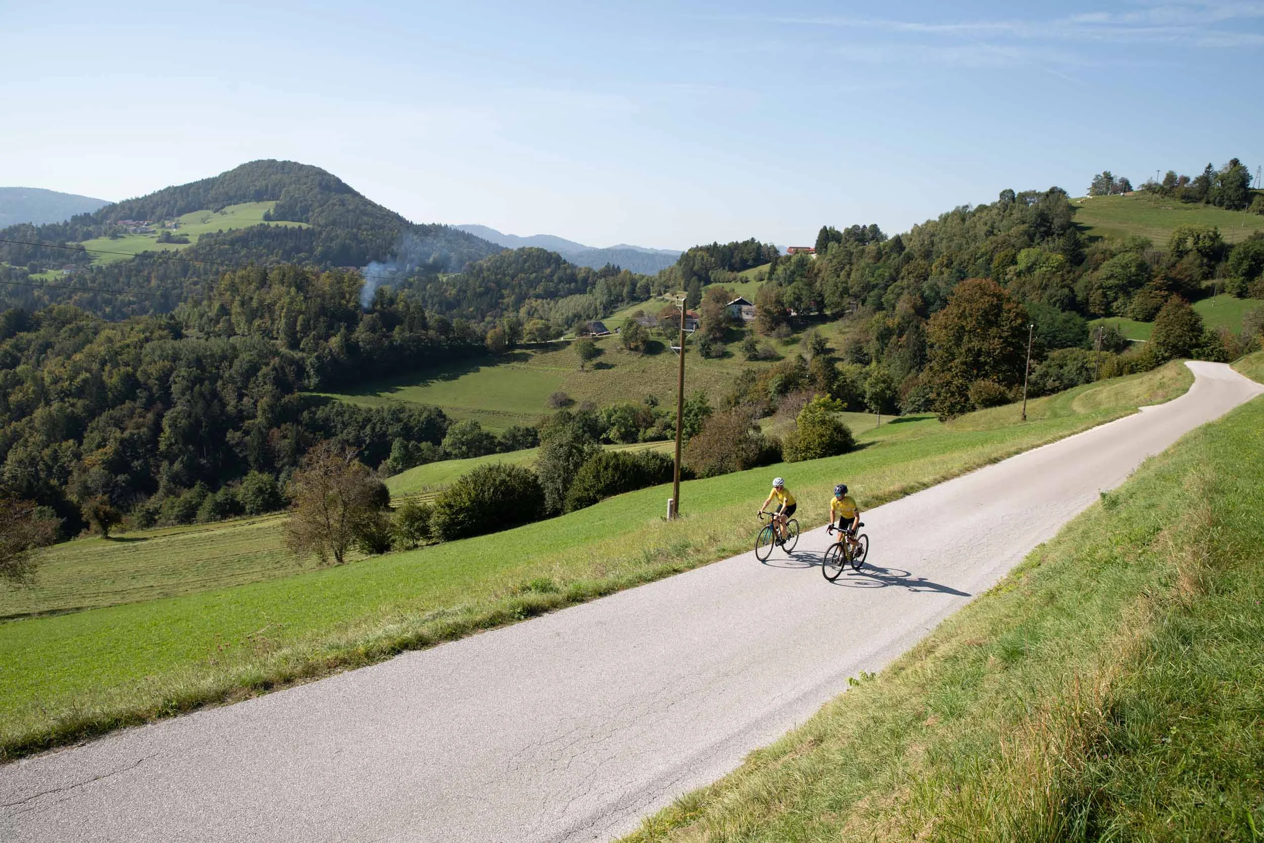 Zwei Radfahrer auf Landstraße mit grüner Hügellandschaft im Hintergrund