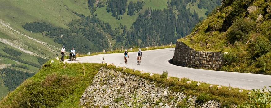 Pinzgau © Heiko Mandl Ciclisti su strada di montagna curva con alberi e nuvole intorno