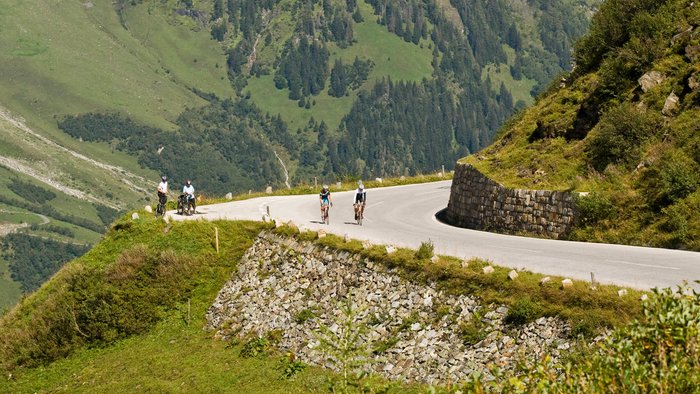 Cyclists riding on a curved mountain road surrounded by greenery and clouds