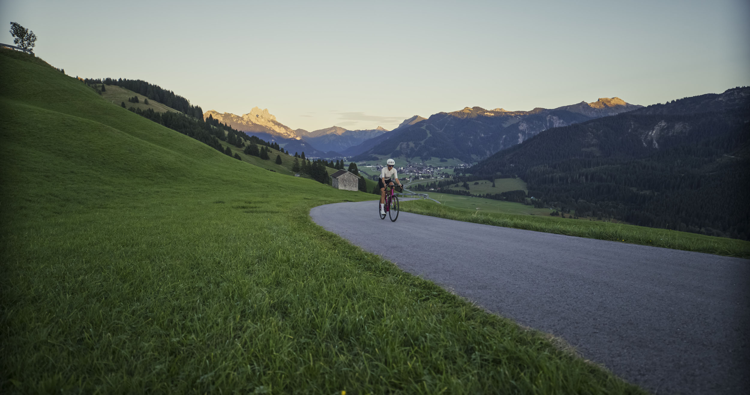 Ciclista su strada di montagna con campi verdi e vista sulle montagne al tramonto