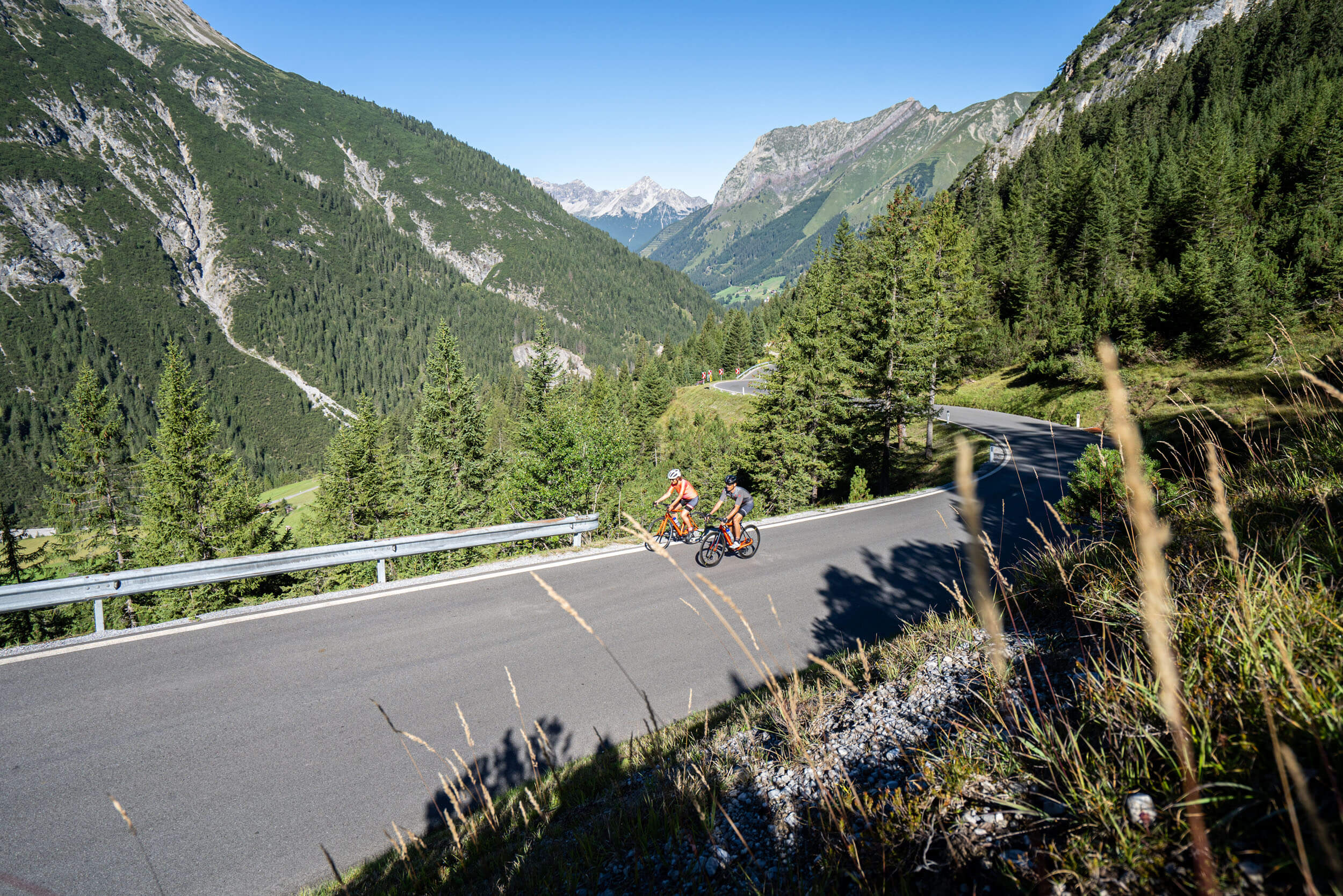 Twee fietsers rijden op bergweg in bebost Alpenlandschap bij heldere lucht
