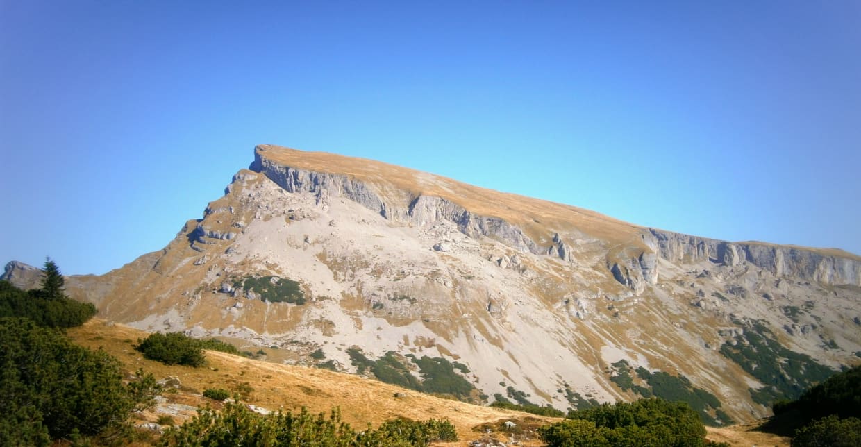 The Mountains are calling Paket Felsiger Berg mit grasbewachsenem Hang unter blauem Himmel