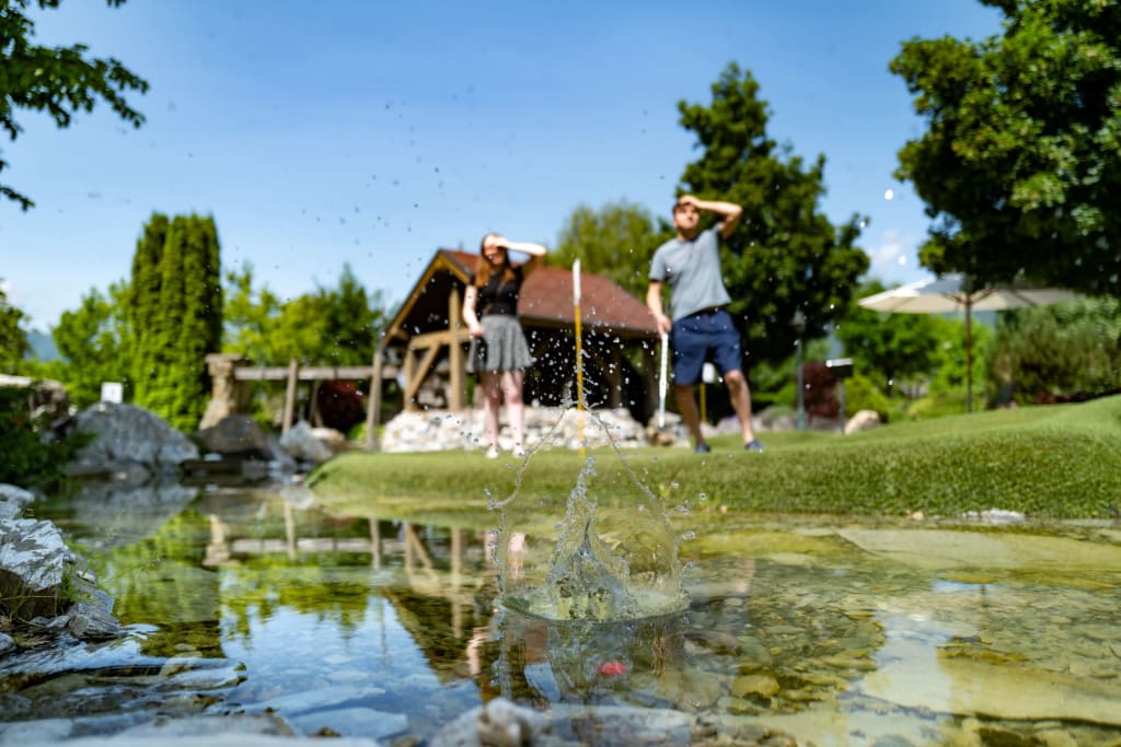 Wasser spritzt in einem Teich beim Minigolf mit zwei Personen im Hintergrund