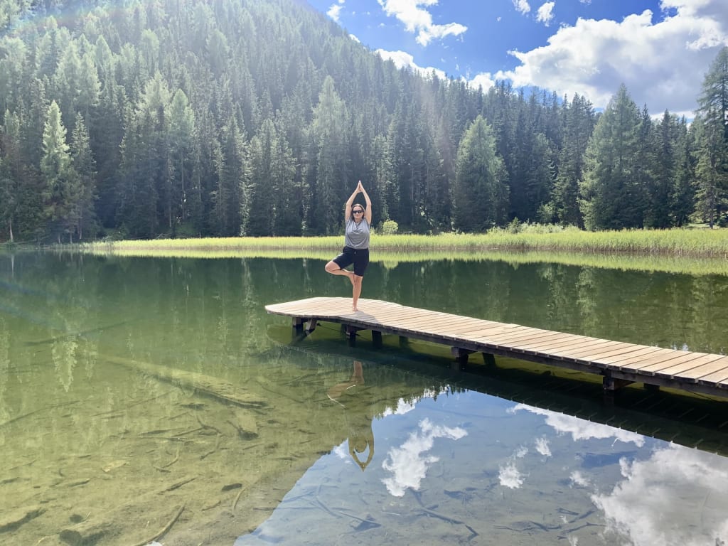 Frau macht Yoga auf Steg über klarem See vor Wald und blauem Himmel