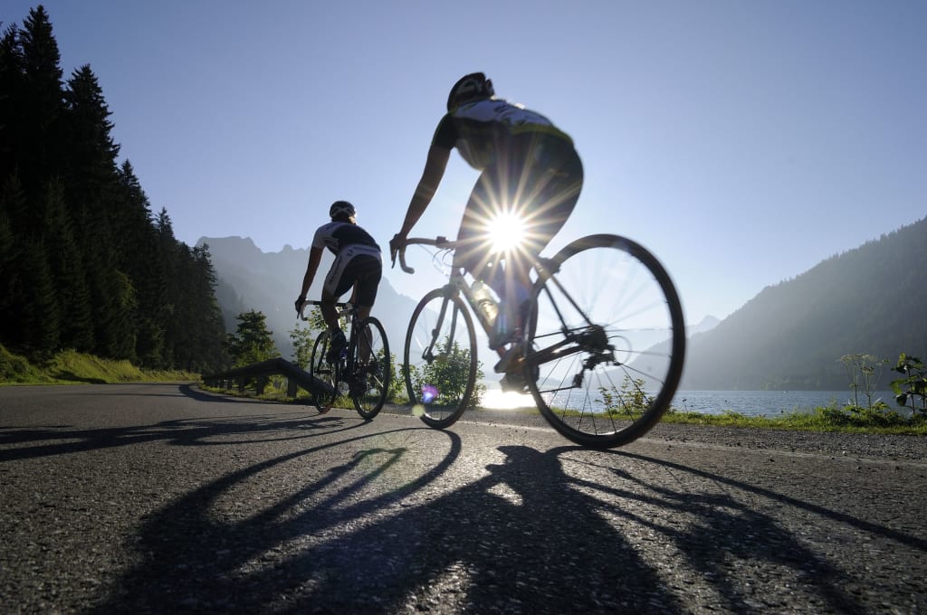 Two cyclists riding on mountain road by lake in sunshine
