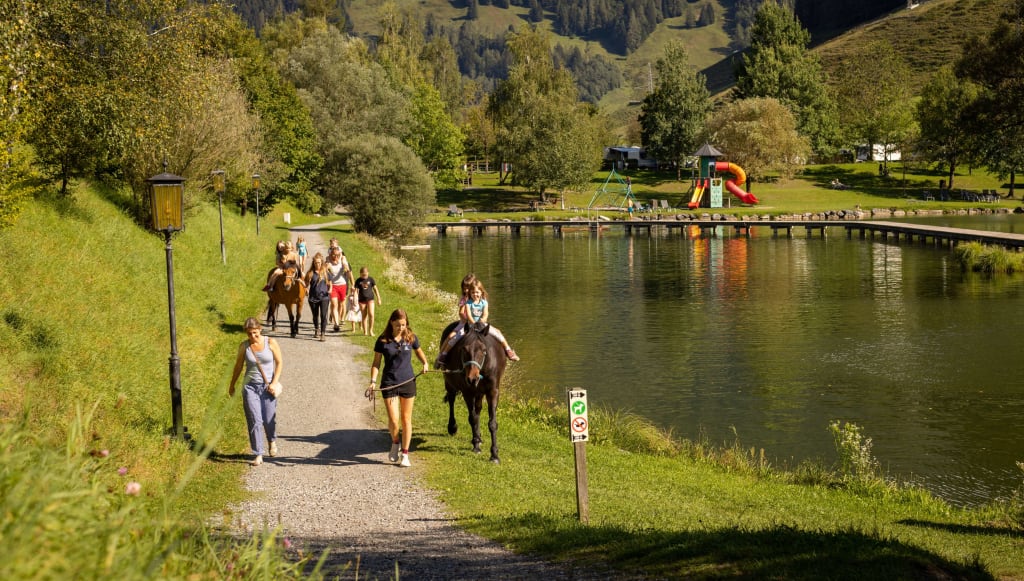 People horseback riding and walking along a lakeside path in a green landscape