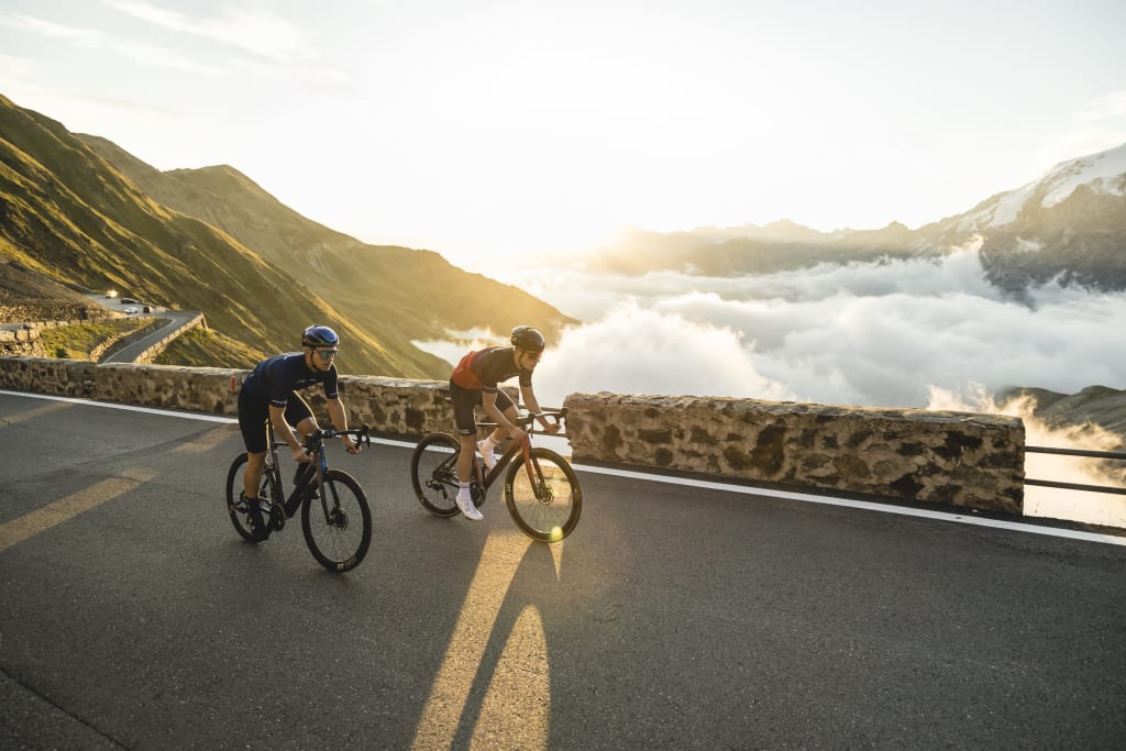 Two cyclists riding on mountain road above clouds at sunrise