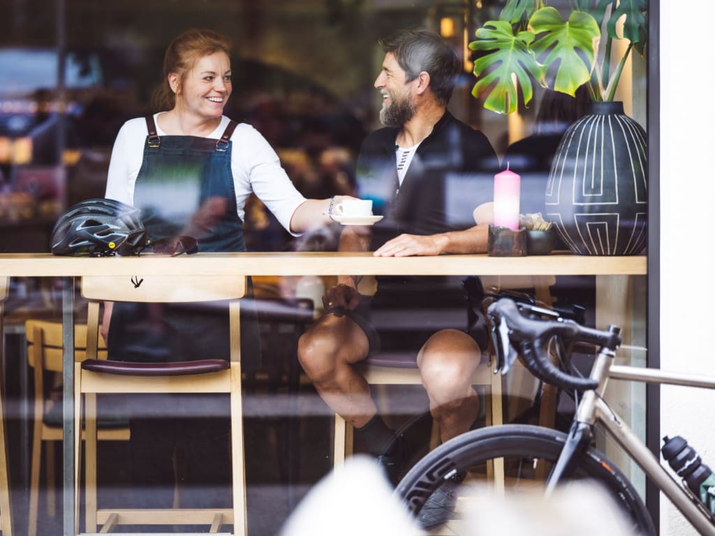 Man and woman laughing and drinking coffee in café, bicycle outside window