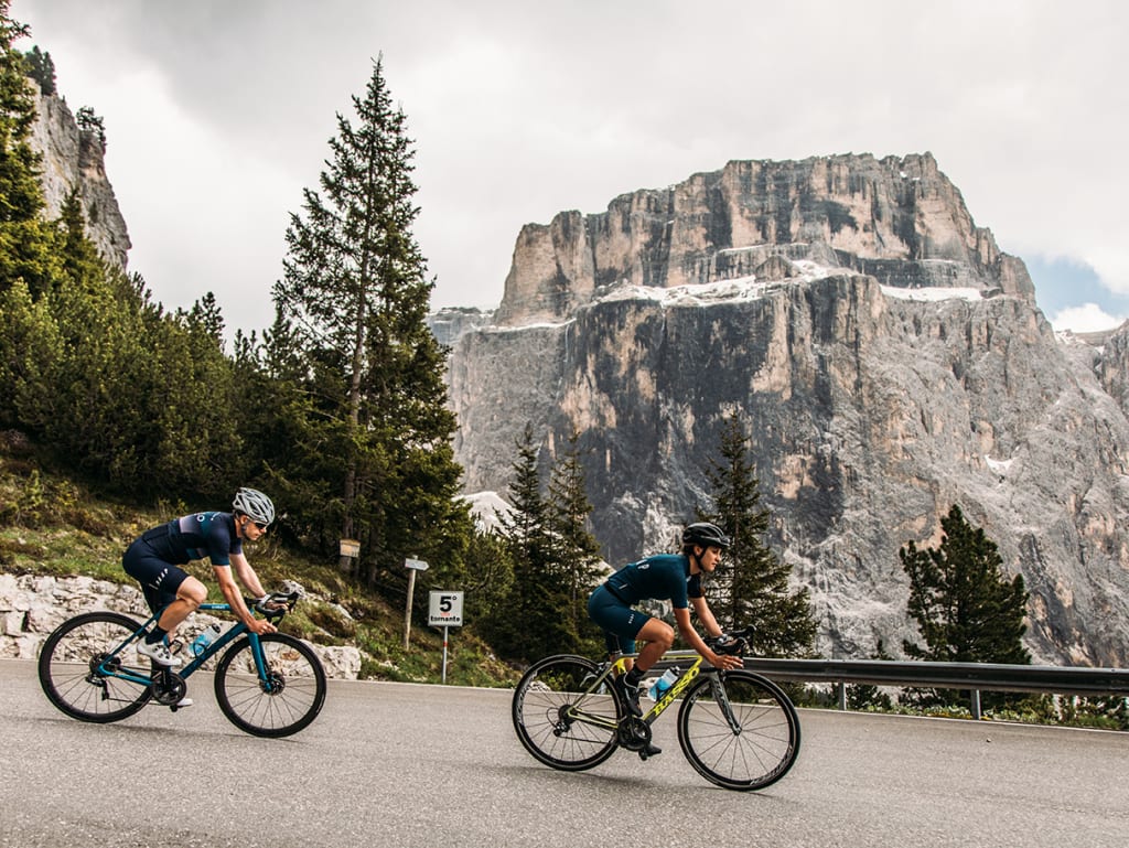 Zwei Radfahrer fahren auf einer Bergstraße mit Felsen und Tannen im Hintergrund