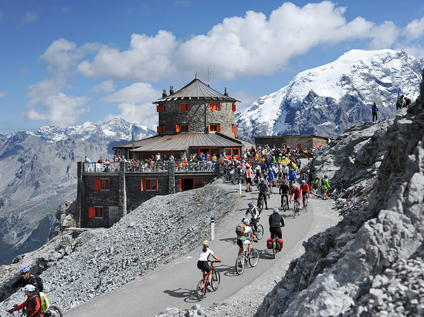 Cyclists on mountain road near mountain lodge with crowd and snowy peak